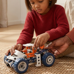 A young child in a red knit sweater sits on a woven rug indoors, playing with a small toy vehicle built from orange, white, and blue building blocks. An adult hand is helping adjust the toy, and a softly lit living room background is visible.