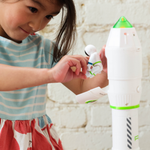 A young girl plays with a white rocket shaped toy that opens to reveal a small astronaut figure, set against a white brick wall.