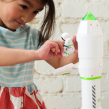 A young girl plays with a white rocket shaped toy that opens to reveal a small astronaut figure, set against a white brick wall.