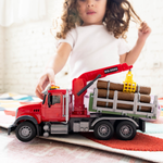 A child's hand and blurred face are visible behind a red toy logging truck carrying logs, placed on a rug in a bright indoor setting.