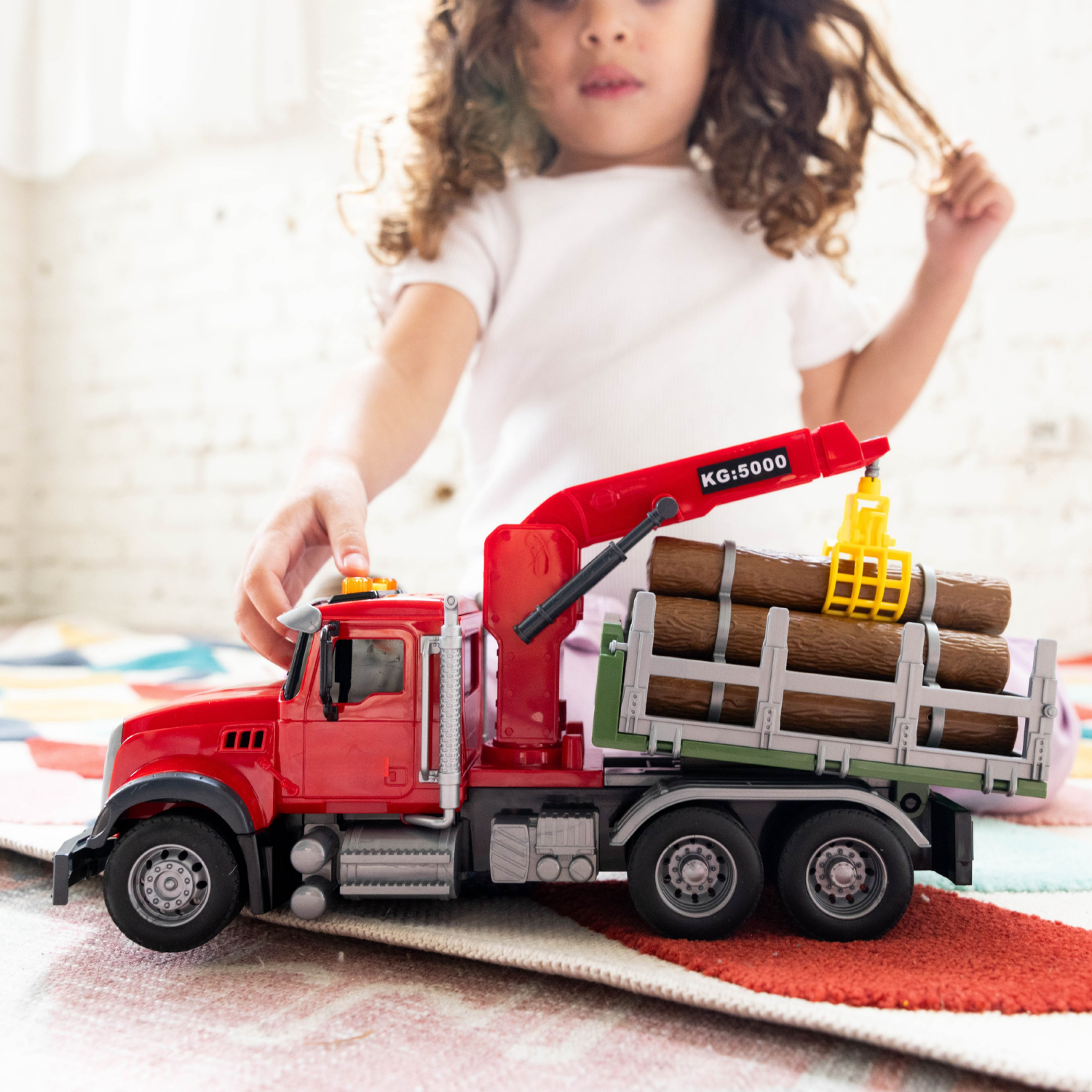 A child's hand and blurred face are visible behind a red toy logging truck carrying logs, placed on a rug in a bright indoor setting.
