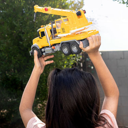 A child viewed from behind holding up a yellow toy crane truck in a backyard with grass, trees, and a gray wall.