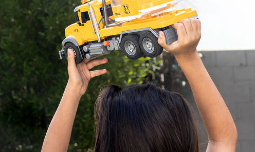 A child viewed from behind holding up a yellow toy crane truck in a backyard with grass, trees, and a gray wall.