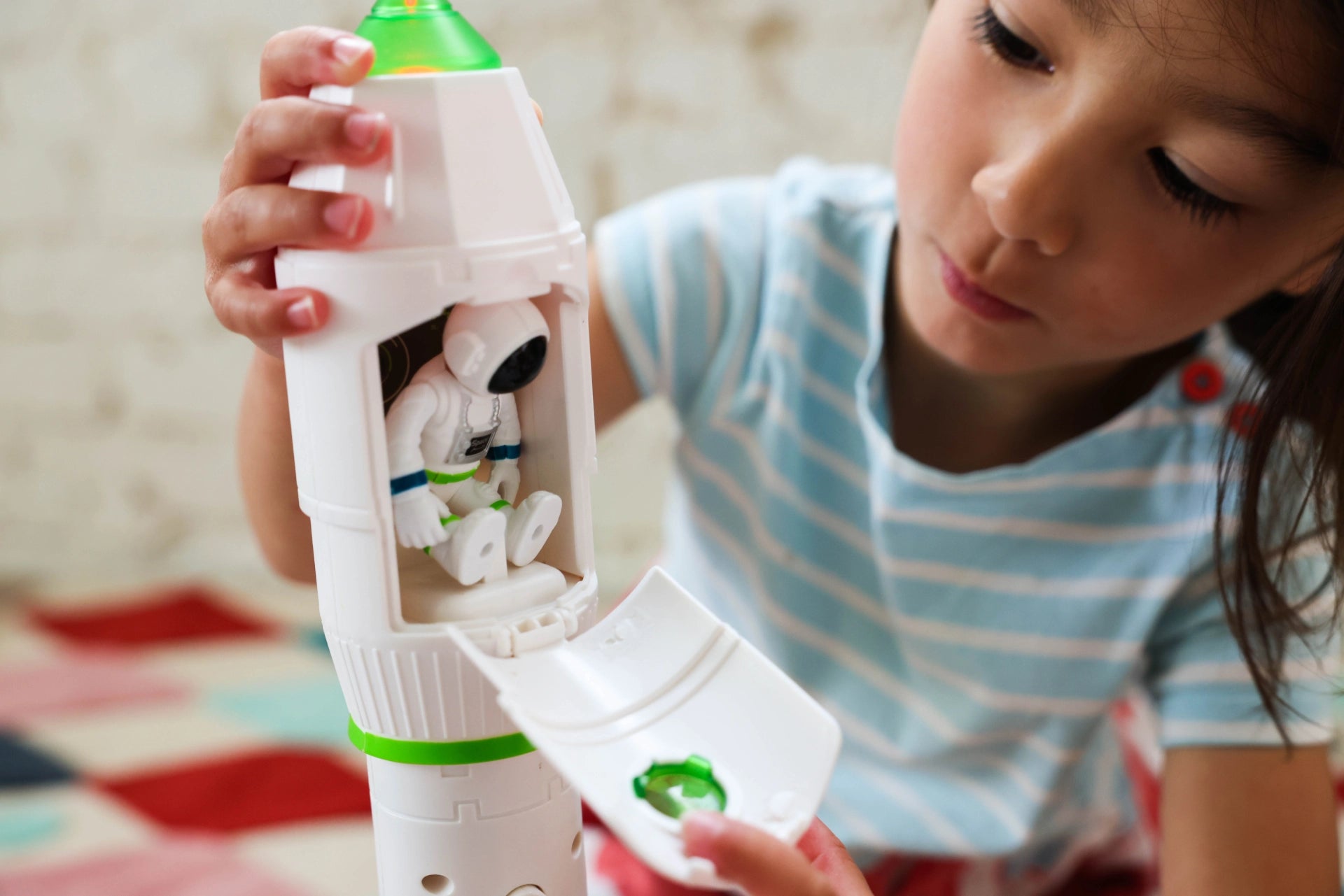 A young child in a light blue and white striped shirt is holding a white toy rocket with a green top and opening a hatch to reveal a small astronaut figure inside.