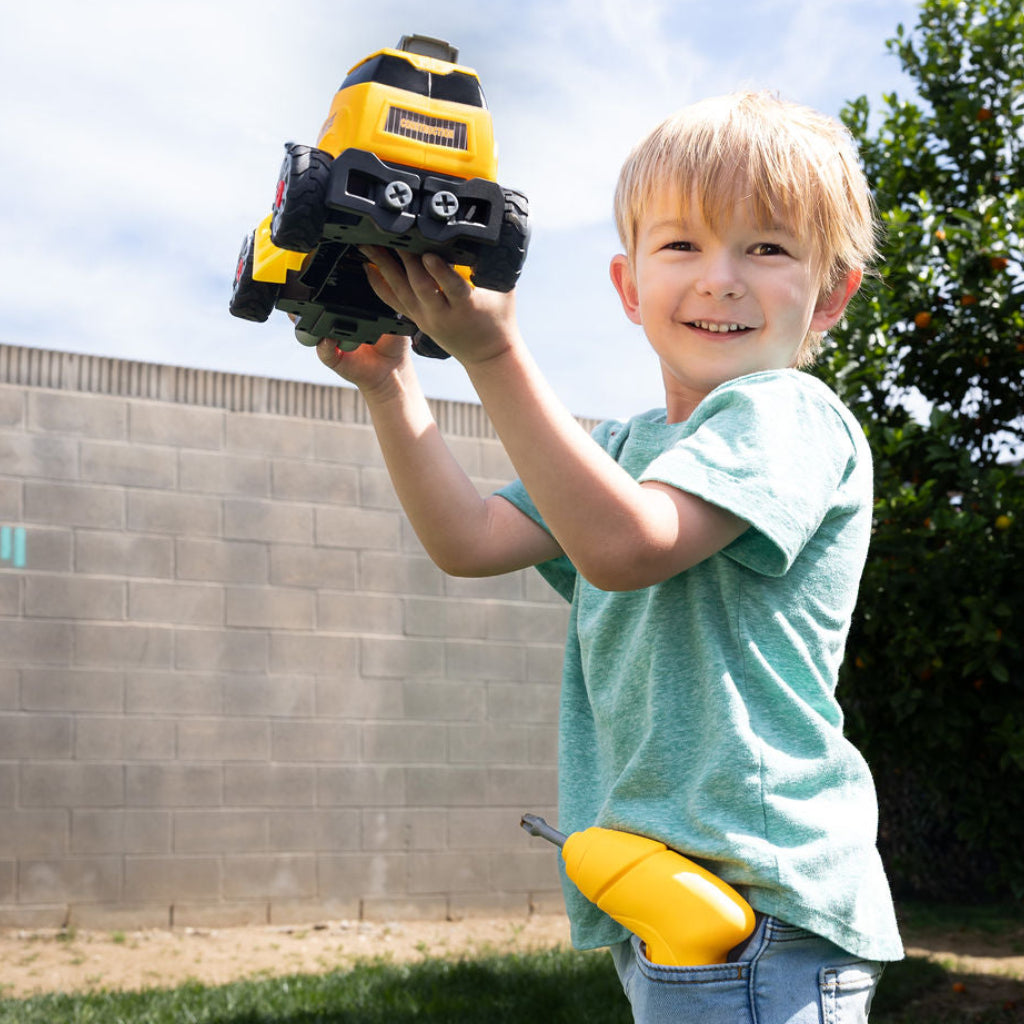 A young boy wearing a light green t-shirt and blue jeans stands outdoors on a grassy lawn, holding a yellow and black toy excavator in his hands. A matching yellow toy is tucked into his waist pocket.