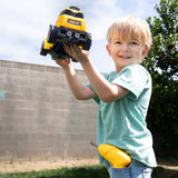A young boy wearing a light green t-shirt and blue jeans stands outdoors on a grassy lawn, holding a yellow and black toy excavator in his hands. A matching yellow toy is tucked into his waist pocket.