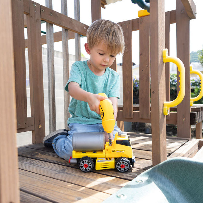 A young boy kneeling on a wooden playground deck, using a yellow toy drill on a yellow construction truck. The scene is set outdoors with green trees and foliage in the background.