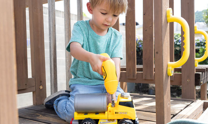 A young boy kneeling on a wooden playground deck, using a yellow toy drill on a yellow construction truck. The scene is set outdoors with green trees and foliage in the background.