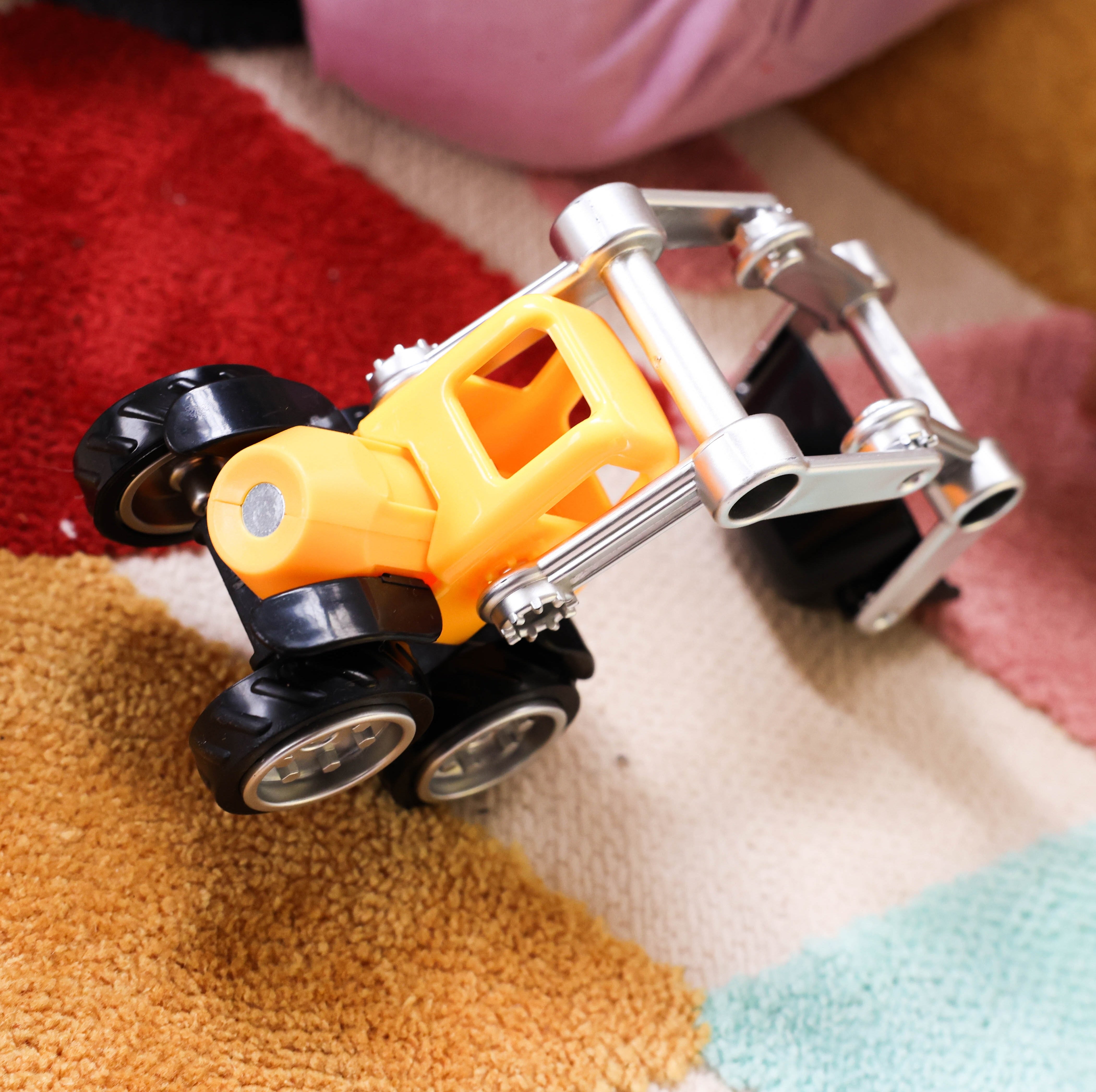 A small toy construction vehicle with a yellow body, black wheels, and a silver metal front loader arm, photographed on a multicolored textured rug.