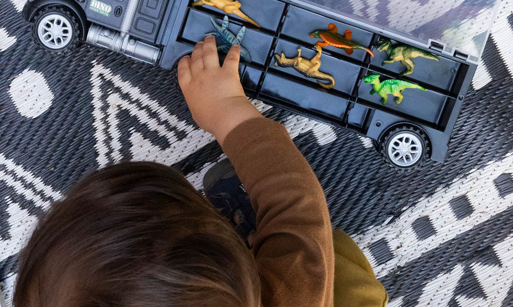 A child's hand and head are visible as they play with a toy dump truck carrying small dinosaur figures on a black and white patterned rug.