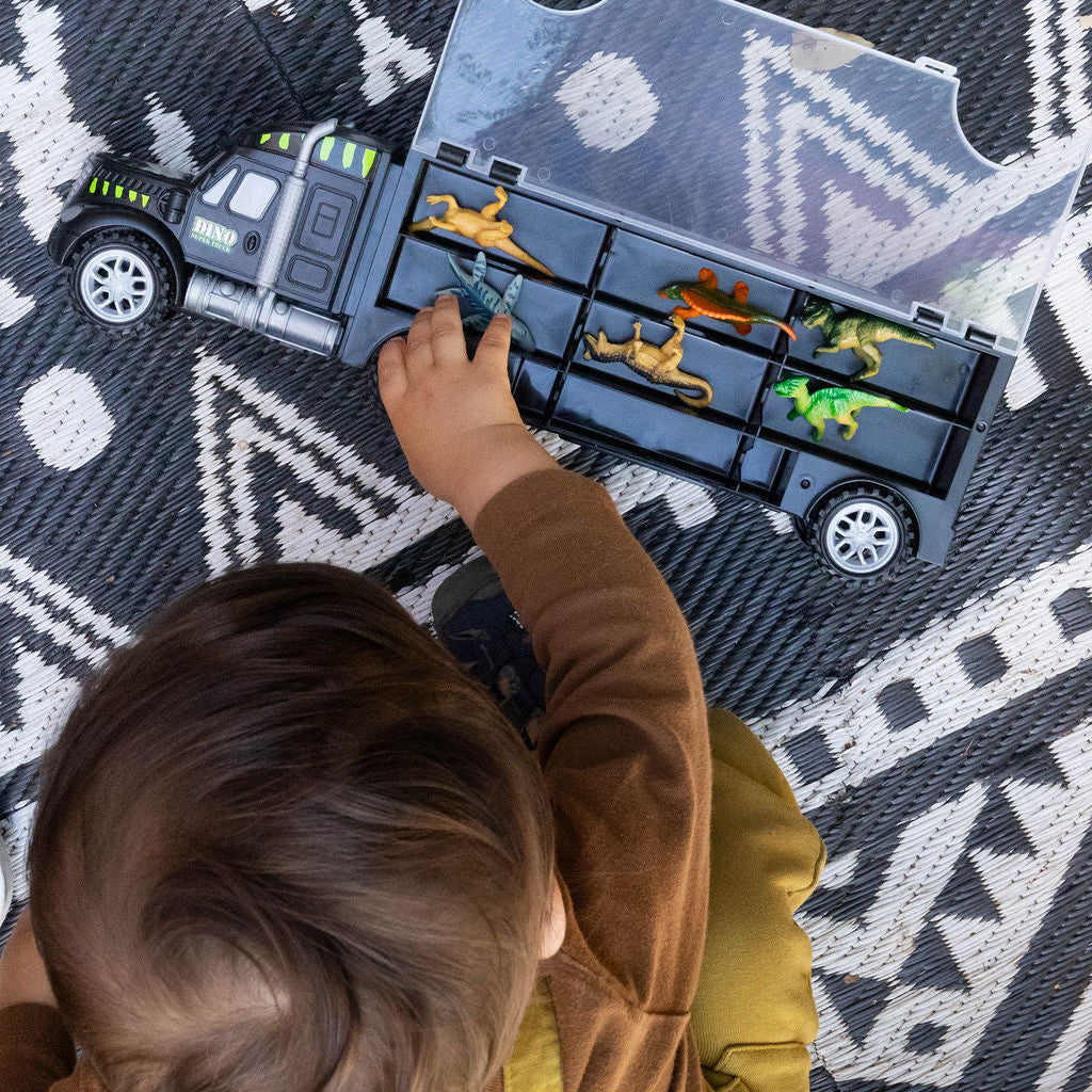A child's hand and head are visible as they play with a toy dump truck carrying small dinosaur figures on a black and white patterned rug.