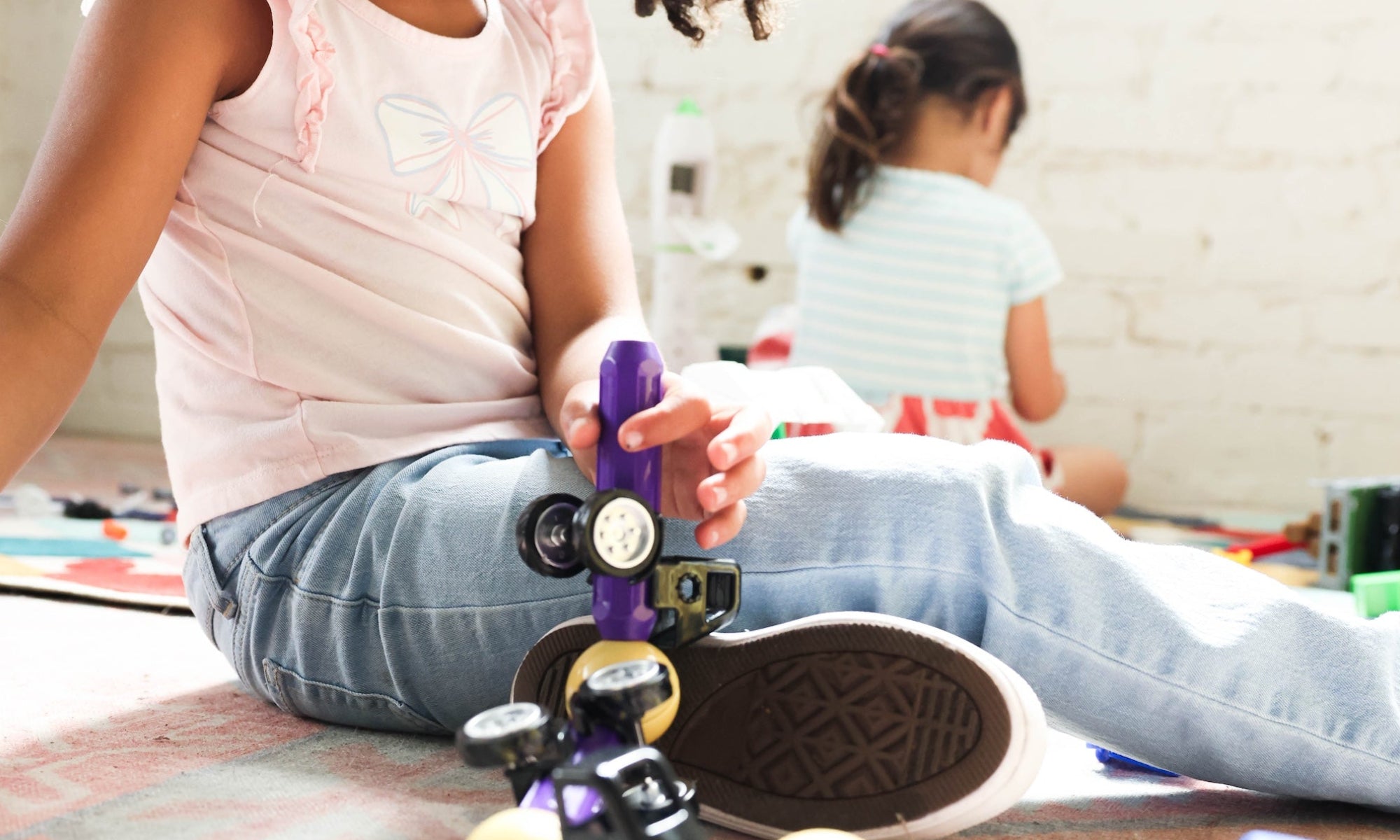 A young girl with curly hair sits on a floor playing with toy vehicles, including a purple and black car and a red and blue truck, in a brightly lit indoor room.
