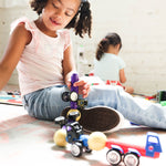 A young girl with curly hair sits on a floor playing with toy vehicles, including a purple and black car and a red and blue truck, in a brightly lit indoor room.