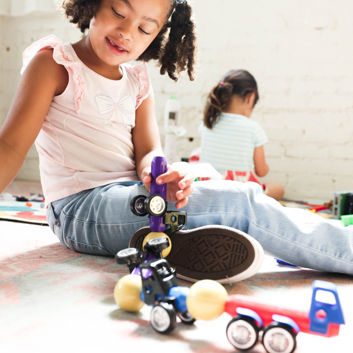 A young girl with curly hair sits on a floor playing with toy vehicles, including a purple and black car and a red and blue truck, in a brightly lit indoor room.