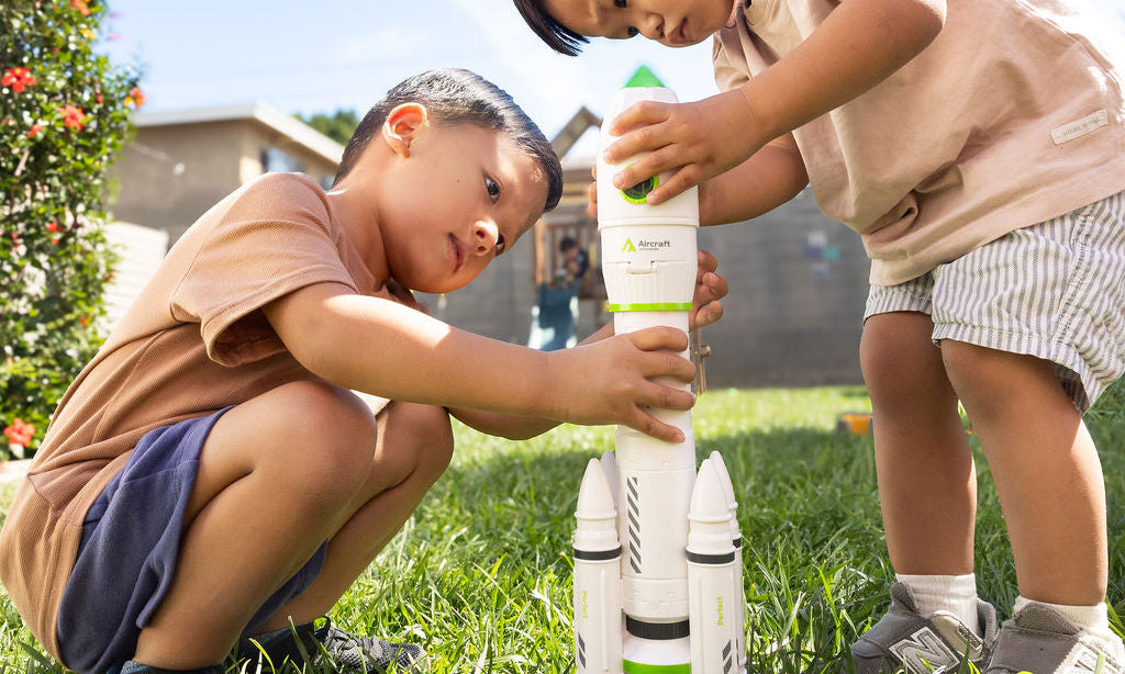 Two children playing with a toy rocketship in a grassy outdoor area.