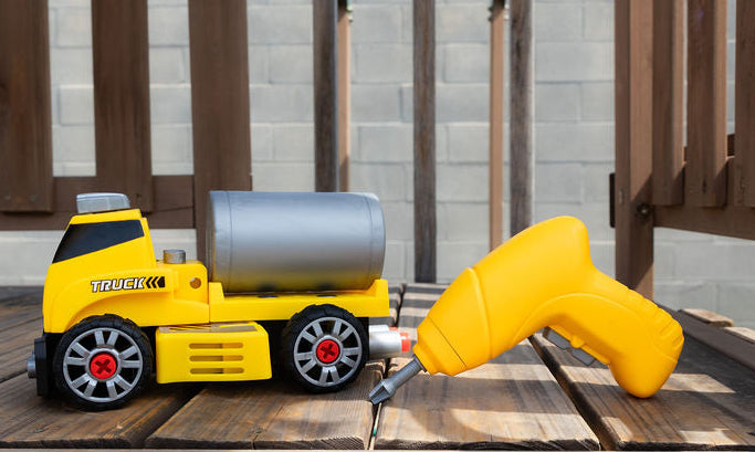 A yellow toy construction truck with a silver cylindrical tank and a yellow handheld drill accessory, placed on a wooden deck in front of a cinderblock wall.