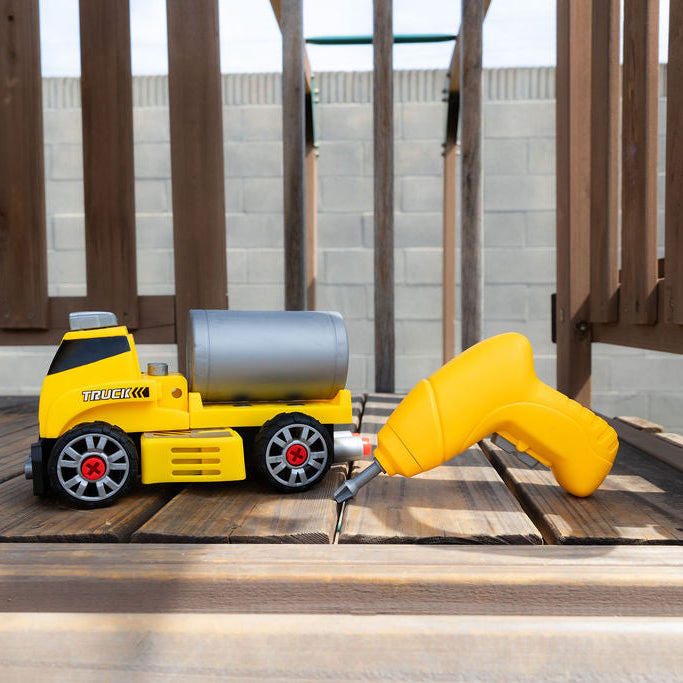 A yellow toy construction truck with a silver cylindrical tank and a yellow handheld drill accessory, placed on a wooden deck in front of a cinderblock wall.