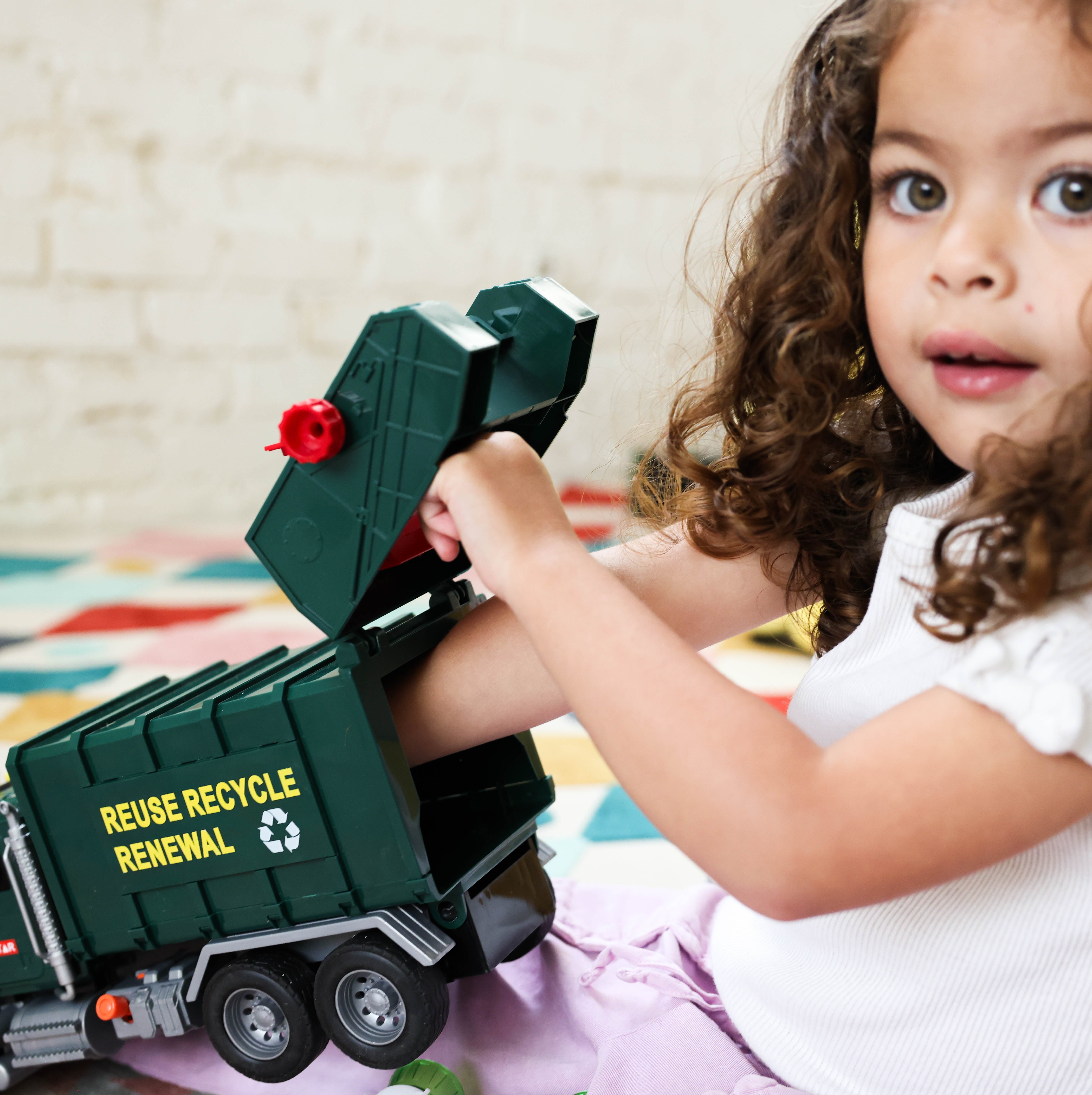 A young child  sits on a patterned rug, playing with a dark green toy garbage truck with her hand inside the back