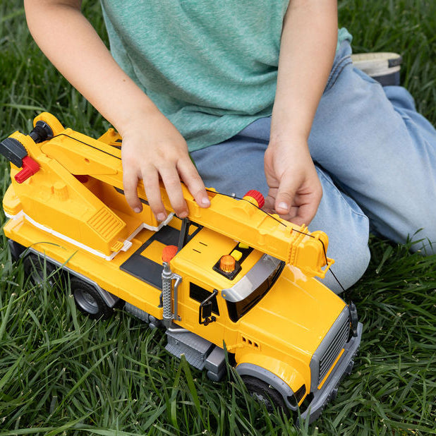 A child kneeling on green grass plays with a yellow toy construction truck with a crane arm.