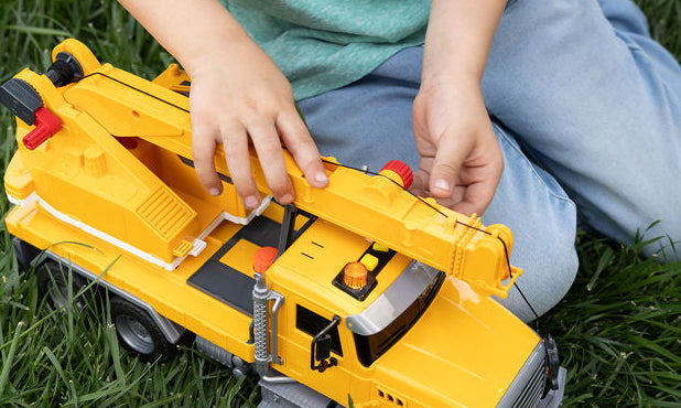 A child kneeling on green grass plays with a yellow toy construction truck with a crane arm.