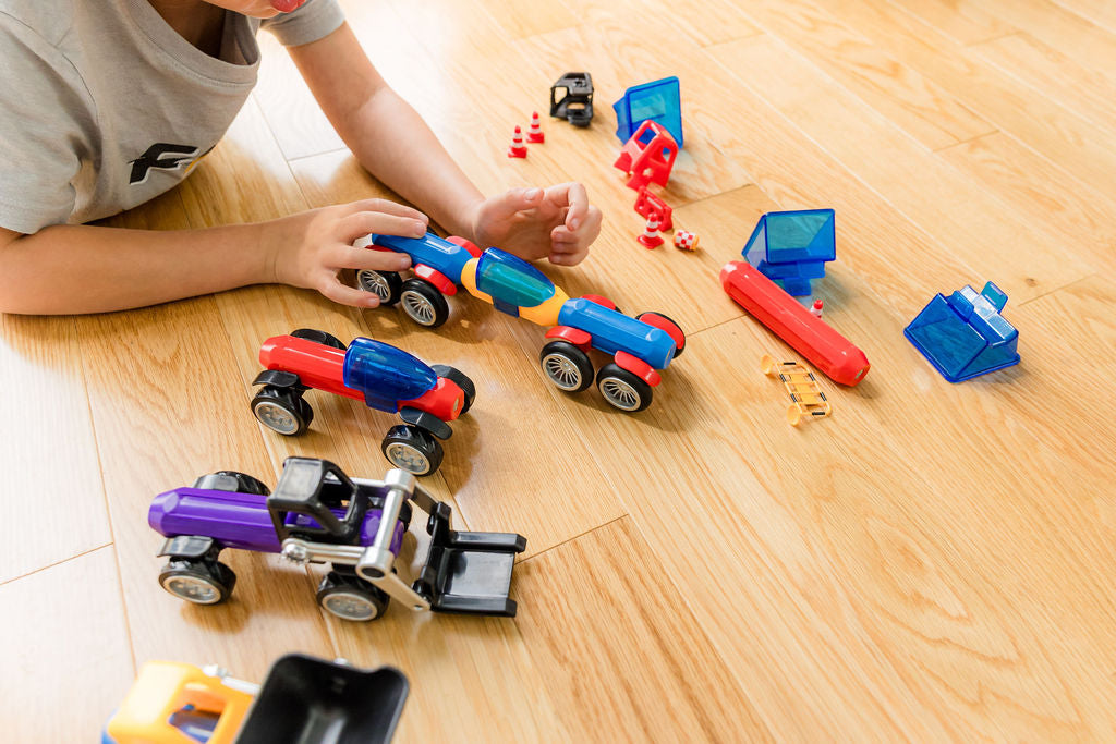 A child's hands are visible as they play with colorful toy construction vehicles and small red building blocks on a light wood floor.