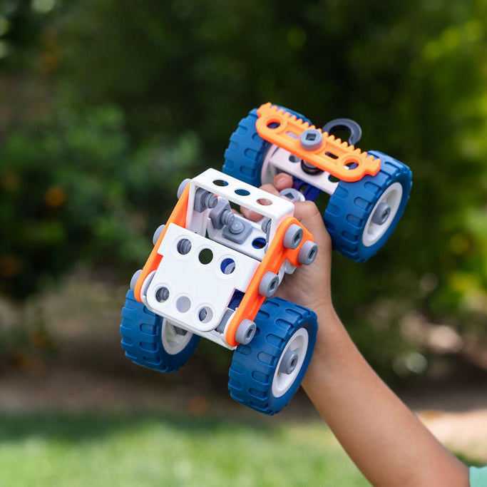 Child holding a small vehicle model made of interlocking bricks against a blurred green outdoor background.
